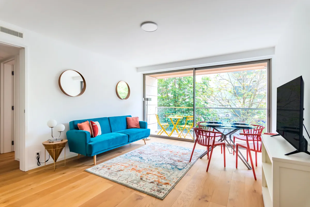 Bright living room with a blue sofa, dining table and a large window onto the balcony in a short-term rental apartment managed by BnBird