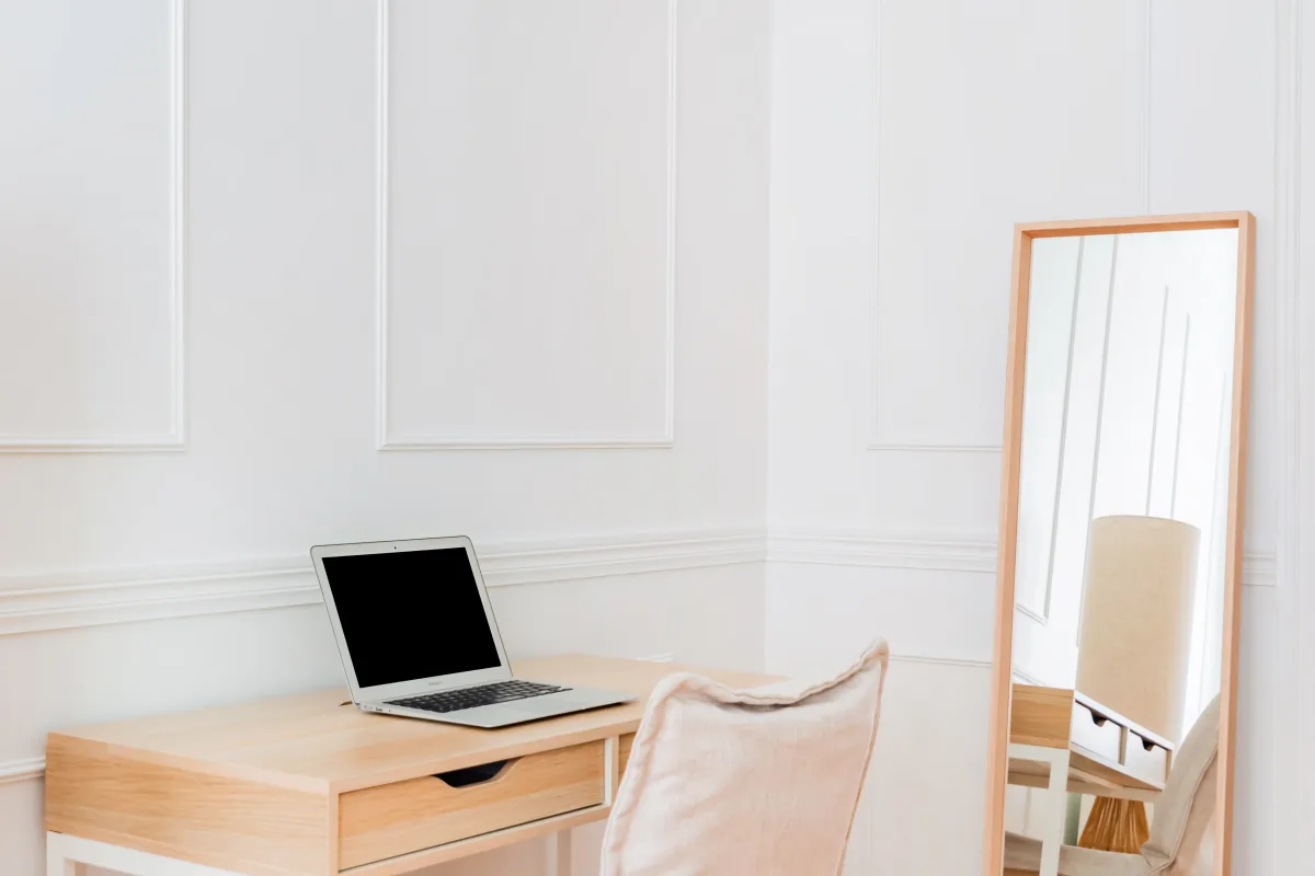 Light wooden desk with a laptop, a cushioned chair and a full-length mirror in a white room